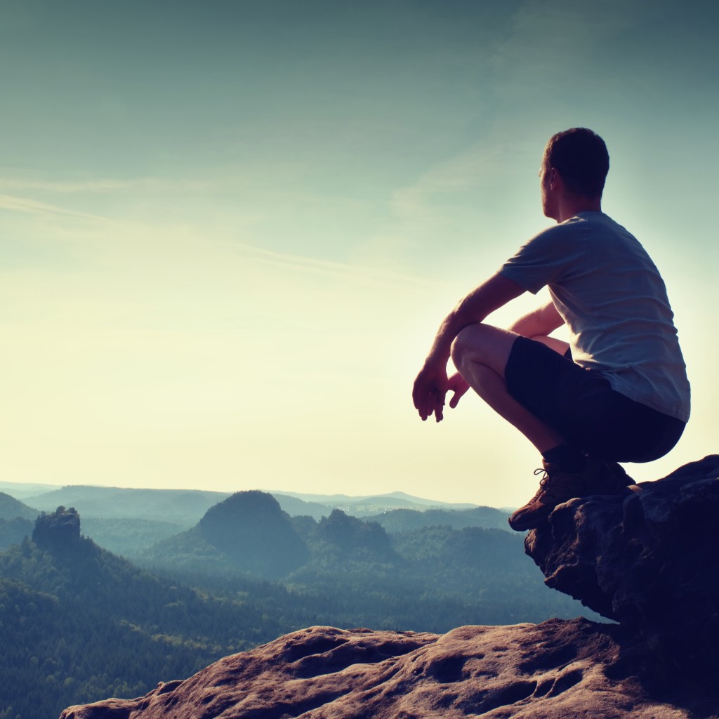 Young man in black sports pants and grey shirt  is sitting on cliff's edge and looking to misty valley bellow